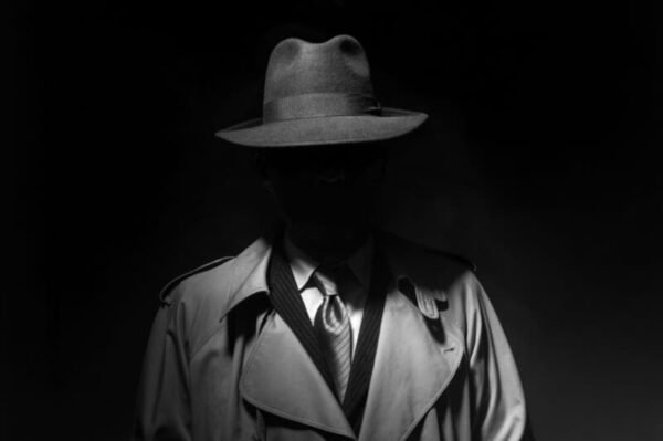 A shadowy figure in a fedora and trench coat stands in a dark, mysterious setting, symbolizing the secrecy and intrigue of private investigation. Learn about Ontario’s private investigator licensing requirements at picourse.ca.